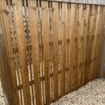 Wooden vertical-slat privacy fence with narrow gaps, set on a gravel-covered ground in a backyard.