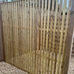 Vertical wooden slat fence panel in a garden, with gravel ground and blue sky above.
