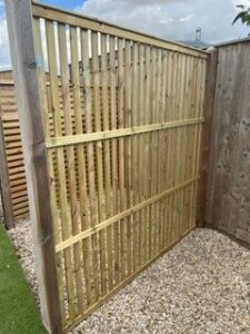 Vertical wooden fence panel with evenly spaced slats leaning against a wooden fence outdoors on a gravel surface.